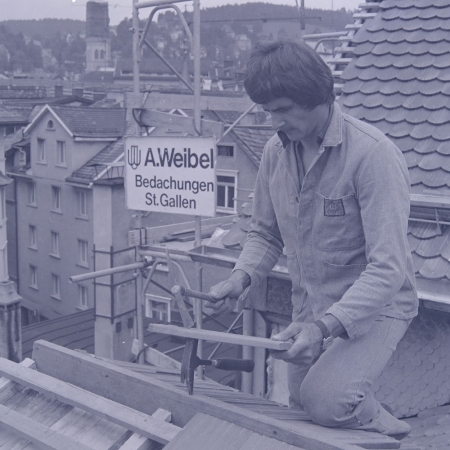 Arbeitsschritte, Werkzeug und nicht zuletzt die Anforderung an die Arbeitssicherheit waren und sind einem steten Wandel unterlegen. Der abgebildete Dachdecker schrotet einen Biberschwanzziegel &ndash; heute w&uuml;rde man hierf&uuml;r die Ziegel- oder Nassfr&auml;se gebrauchen. Dachdecker-Meister-Verband, 1978. StadtASG, PA Foto Gross, TA50257.
